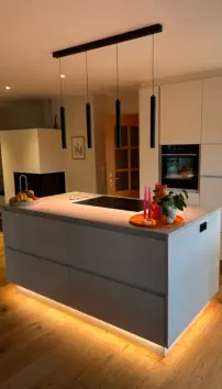 Graue Kochinsel mit Kochfeld und warm leuchtender LED-Sockelbeleuchtung unter schwarzen Pendelleuchten