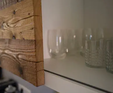 Detail einer Blende aus Altholz mit handgehackter Oberfläche neben einem weißen Regal mit Gläsern