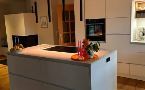 Küche nach Maß mit grauer Kochinsel, warm leuchtender LED-Sockelbeleuchtung und hellen Hochschränken
