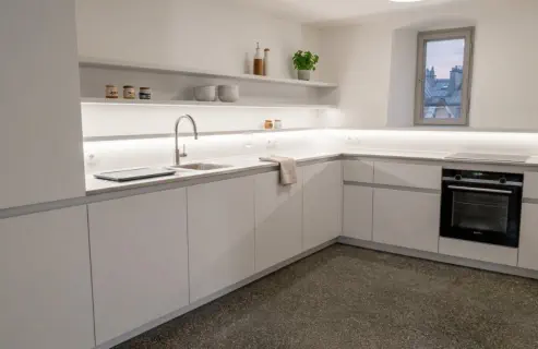 Weiße grifflose Küche in L-Form mit beleuchtetem Wandbord, eingebautem Backofen und heller Arbeitsplatte