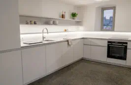 Weiße grifflose Küche in L-Form mit beleuchtetem Wandbord, eingebautem Backofen und heller Arbeitsplatte
