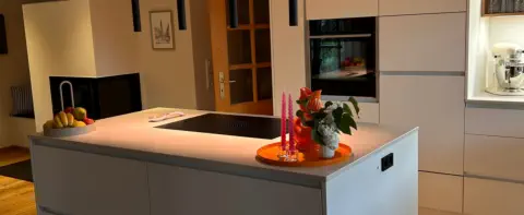 Küche nach Maß mit grauer Kochinsel, warm leuchtender LED-Sockelbeleuchtung und hellen Hochschränken