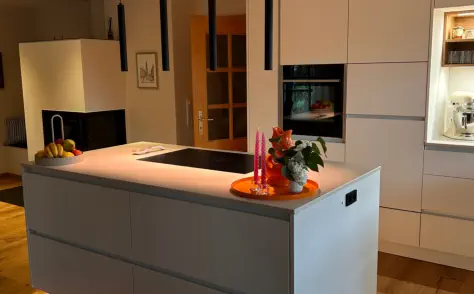 Küche nach Maß mit grauer Kochinsel, warm leuchtender LED-Sockelbeleuchtung und hellen Hochschränken
