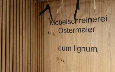 Schriftzug Möbelschreinerei Ostermaier cum lignum in Eichenholz neben einer Lamellenwand